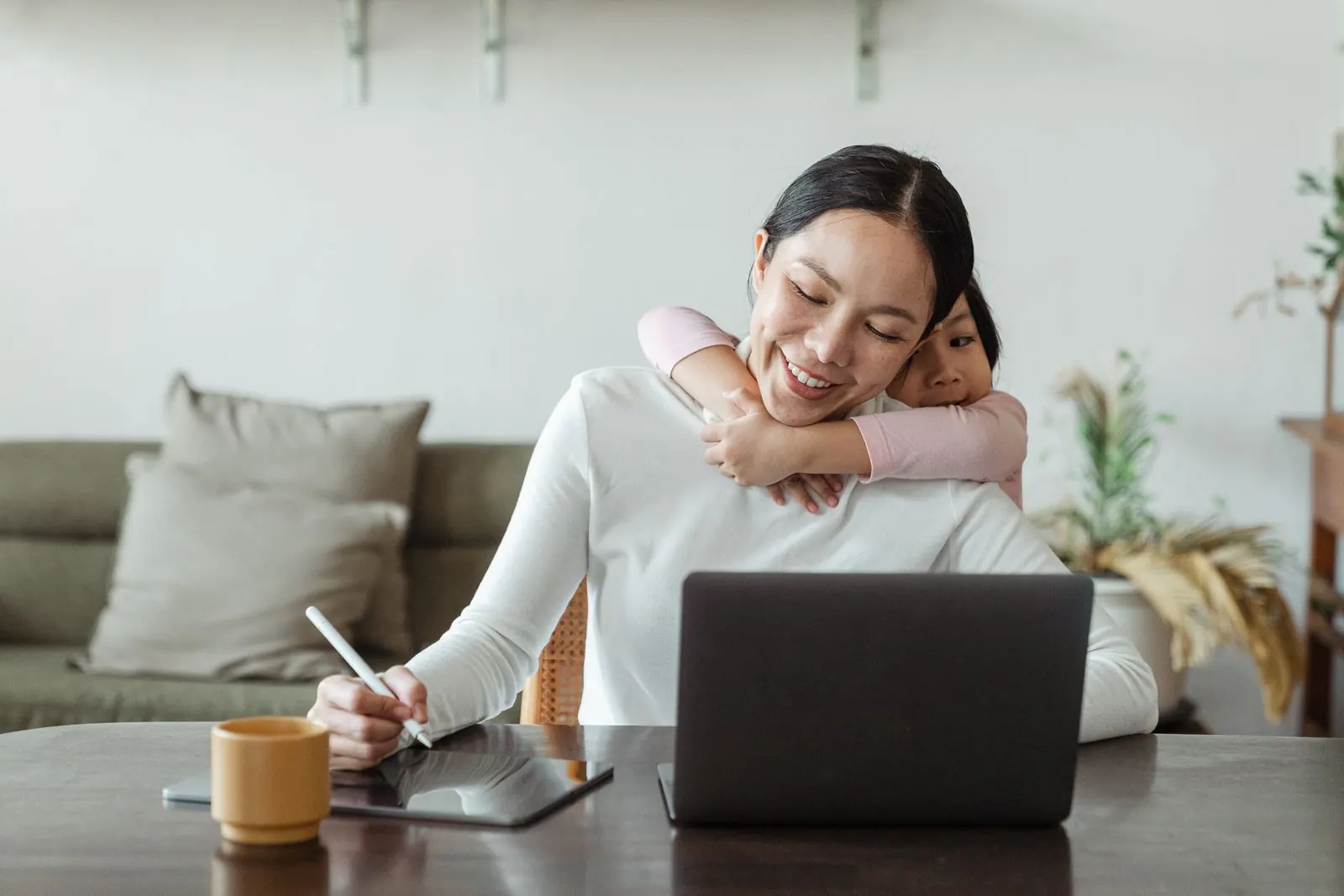 Người mẹ ghi chép bên bàn, con gái ôm mẹ từ phía sau